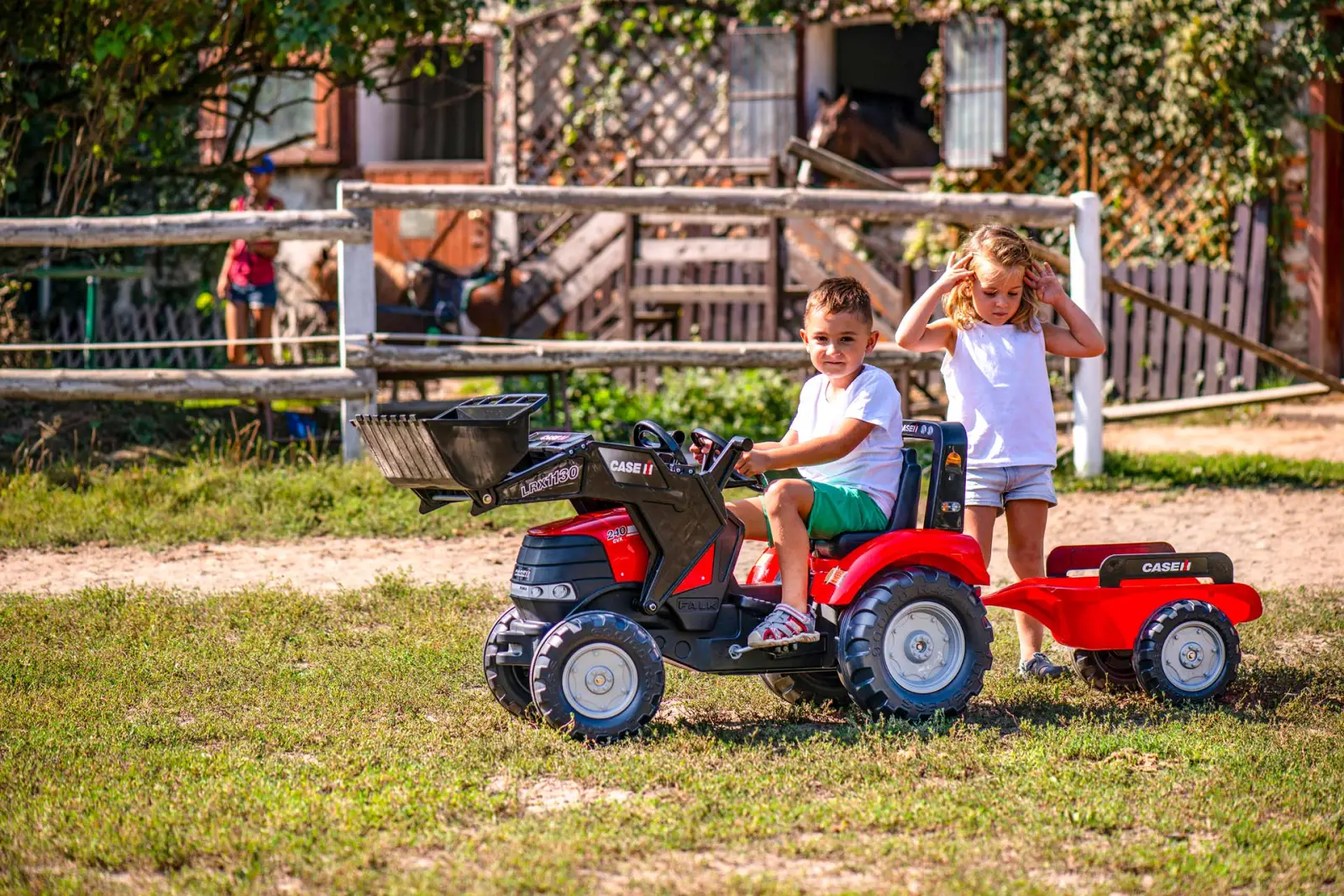 Deux enfants dans une ferme, l’un au volant d’un tracteur à pédales Falk Case IH attelé à sa remorque.